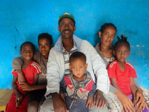 A husband and wife sit with their four children next to them and on their laps, all smiling for the camera