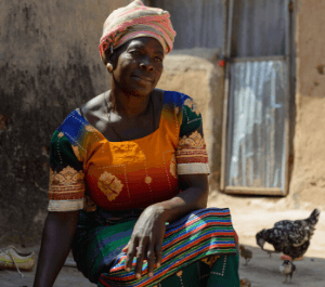 A woman sits in front of her house with a chicken next to her