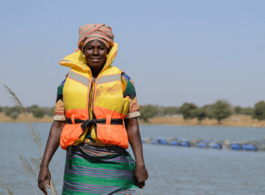 A woman stands in a lifejacket in front of the shoreline, smiling for the camera