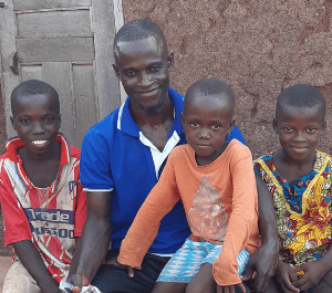 A man sits in front of his house with his three children around him
