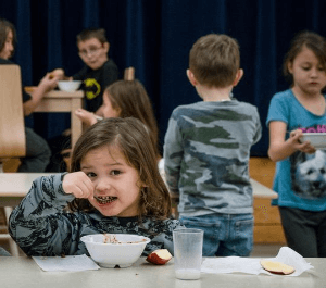 Close up of a young child eating a bowl of cereal with other children eating in the background
