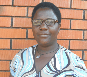 A woman stands in front of a brick wall, smiling for the camera