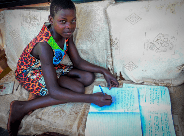 A young girl sits on her couch doing her school work, smiling for the camera