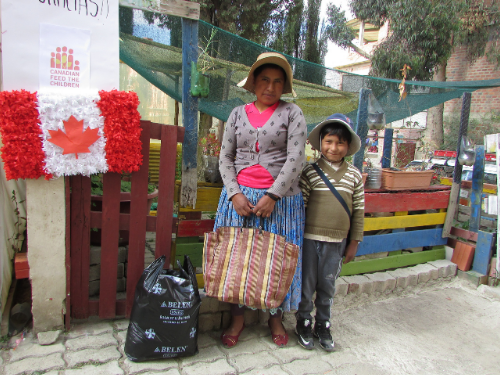 A mother and son stand in front of a fence smiling with food hampers