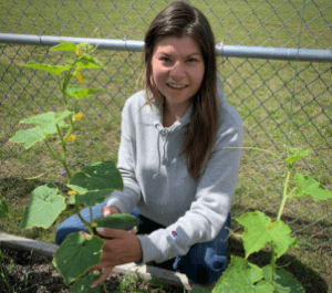 Kiana wears a grey hoodie while crouching behind a cucumber plant. She holds the cucumber in her hand while smiling facing the camera. Behind her is a chainlink fence.