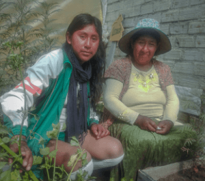 Jhoselin at left crouches next to her mother in the urban garden in Bolivia. They are surrounded by a brick wall and plants grow around them.