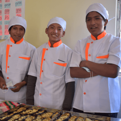 Three boys in cooking uniforms stand in front of a tray of food they prepared.