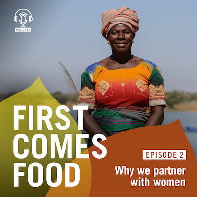 First Comes Food: Episode 2 Why we partner with women. Talata stands tall in a beautiful Ghanaian dress and headwrap, smiling confidently against a backdrop of the river where she farms her fish, and a cloudless blue sky.