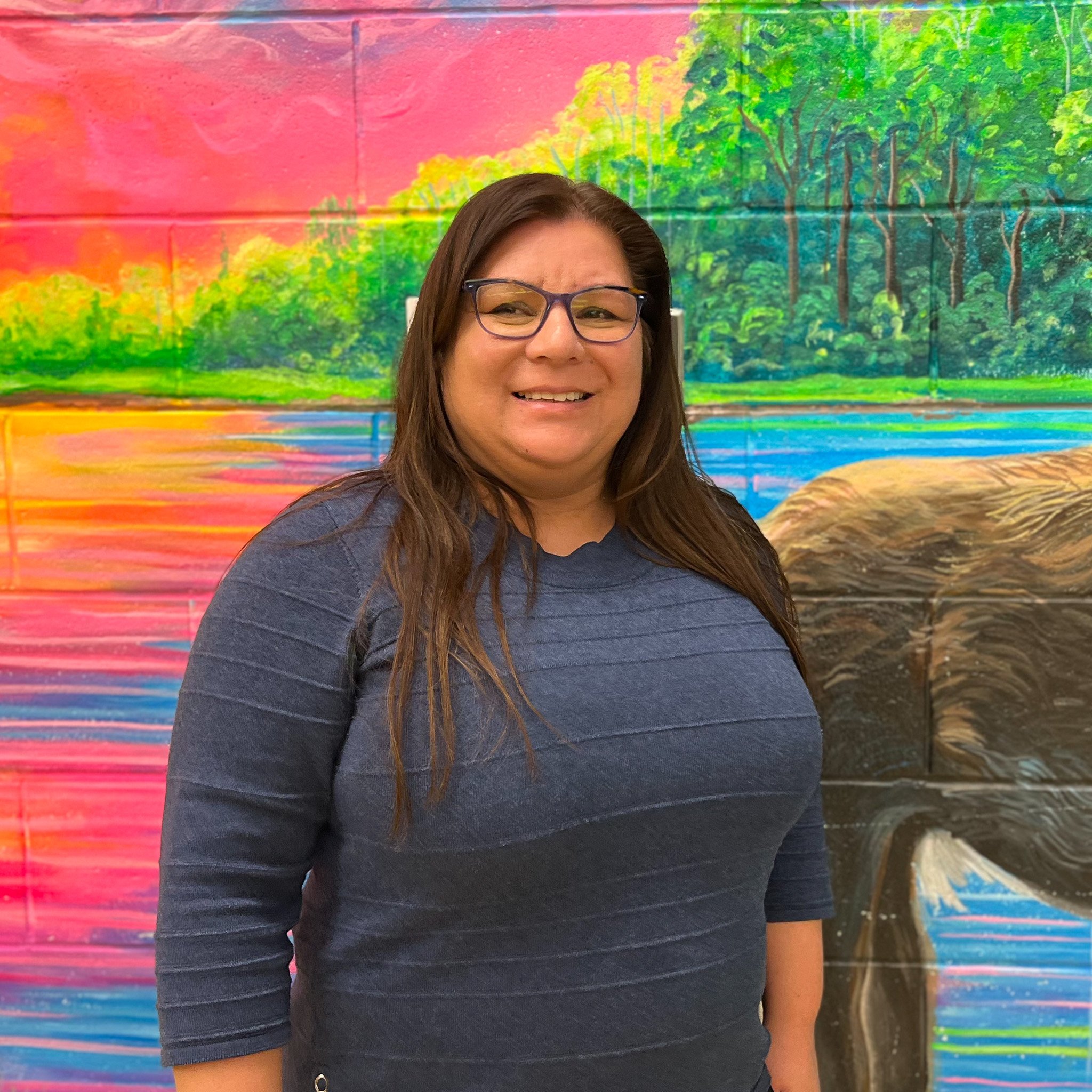 Terri-Anne smiles in front of a brightly painted mural of a river at sunset inside of the school. The greenhouse will provide fresh produce for the school meal program.