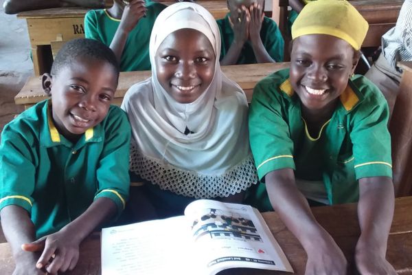 Three Ghanaian children in green shirts sit at a desk in a classroom, smiling for the camera