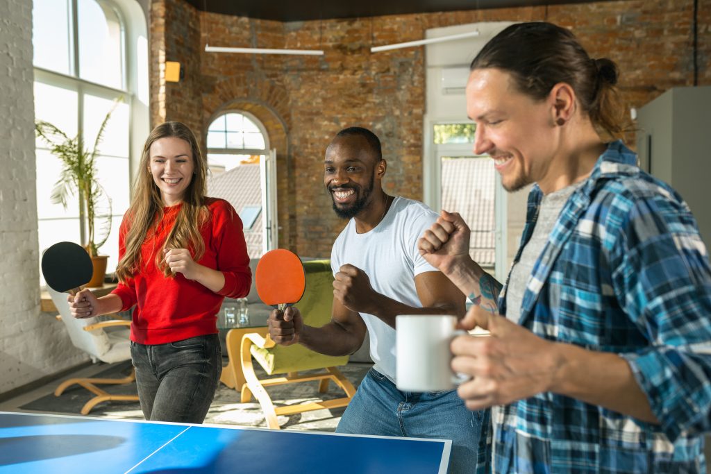 Young people playing table tennis in workplace, having fun. Friends in casual clothes play ping pong together at sunny day. Concept of leisure activity, sport, friendship, teambuilding, teamwork.