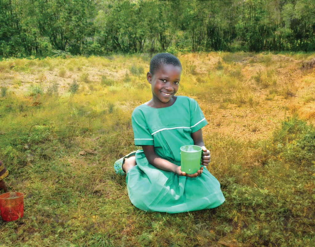 a girl sits in the grass, smiling, holding a cup of food