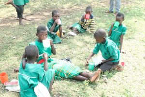 a group of school children sit in the grass, eating breakfast together