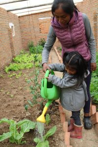 a woman stands smiling over her daughter using a watering can in the garden